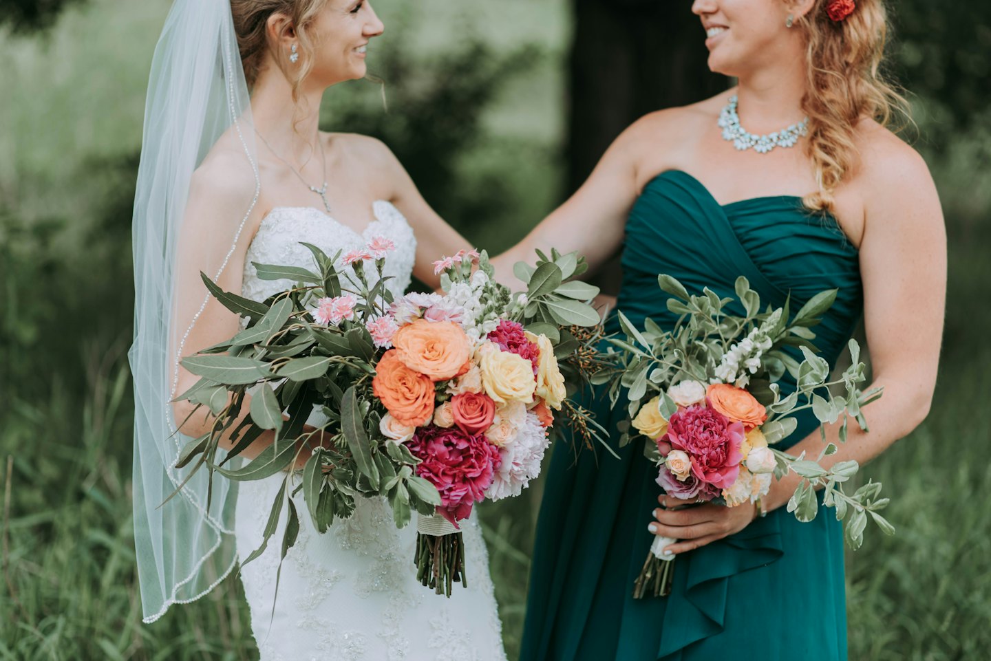 Bride and bridesmaid smile at each other holding bouquets.