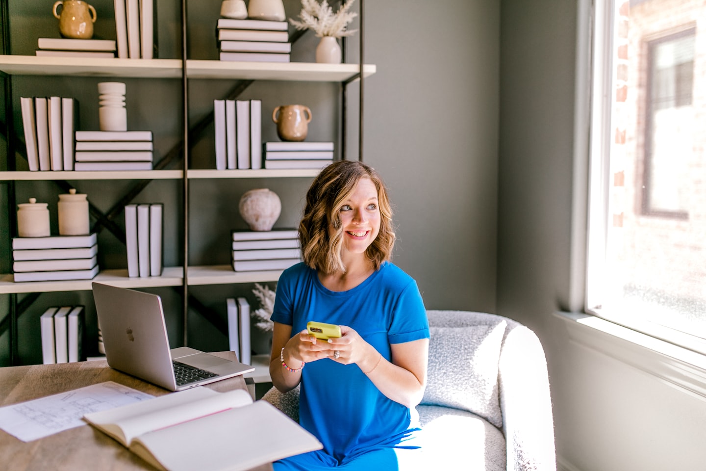 Woman in blue dress uses smartphone at desk with laptop and bookshelf.