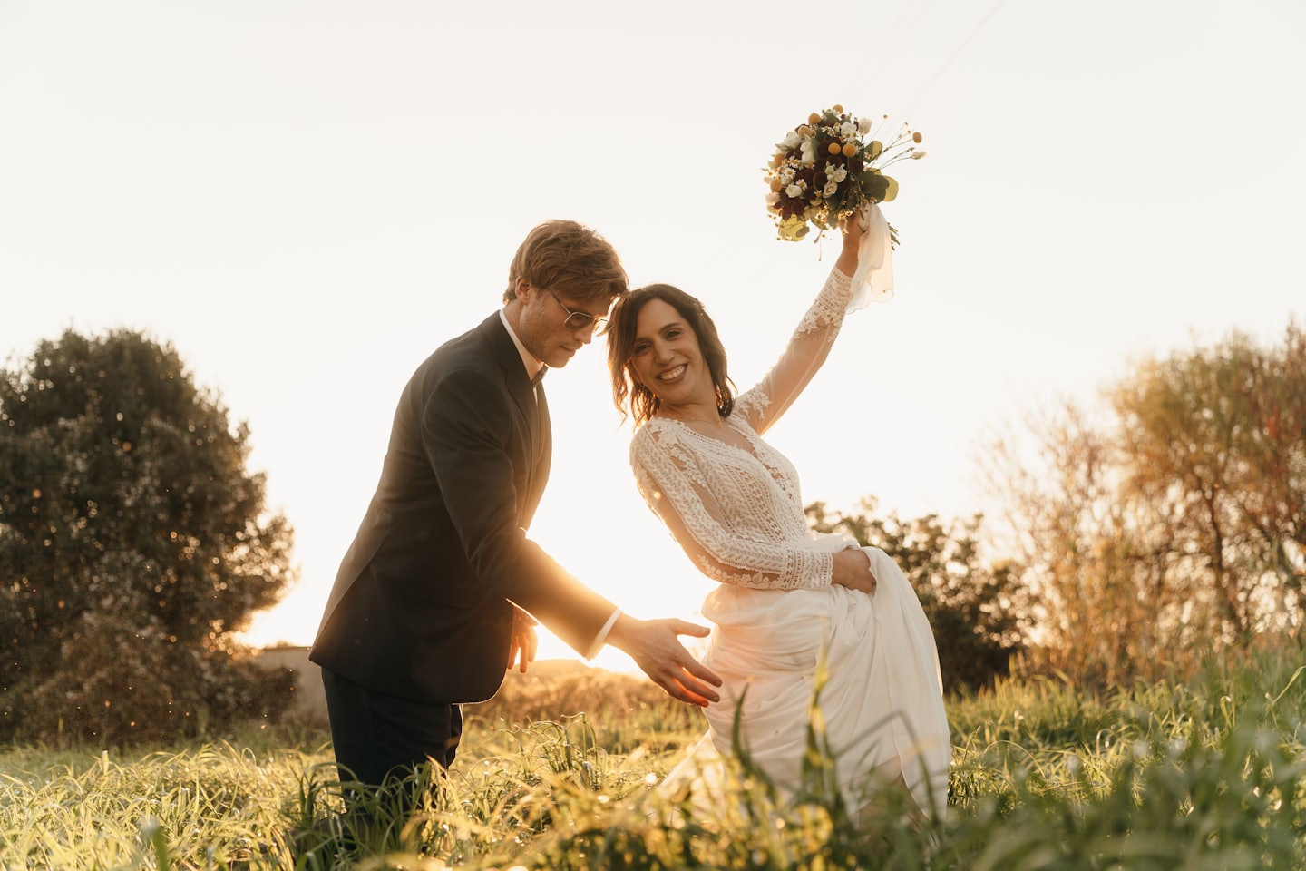 Newlyweds stand in a sunny field with a wildflower bouquet.