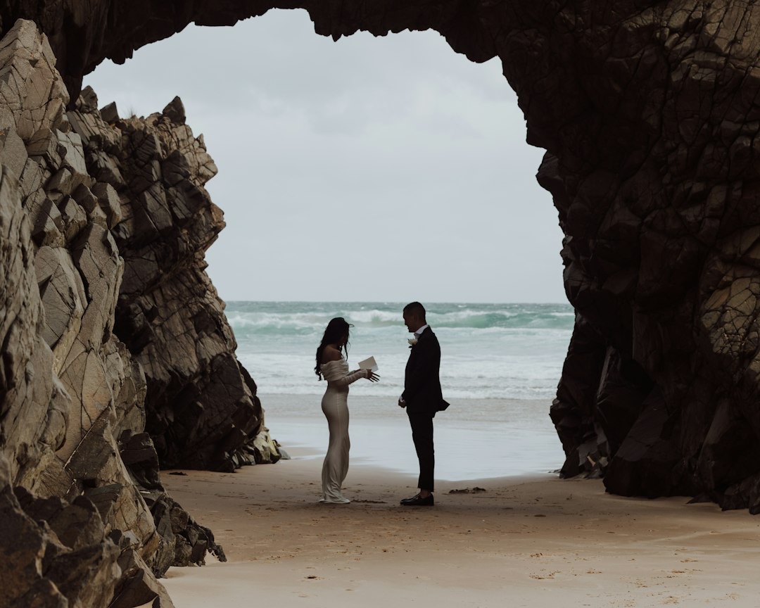 Couple on beach reading vows under rock arch.
